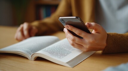 Fototapeta premium A young Asian woman holds a smartphone while reading a book, immersed in a cozy indoor setting.