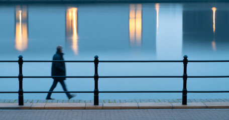 Blurred silhouette of a person walking along a waterfront promenade with metal railings and glowing reflections on calm water at dusk
