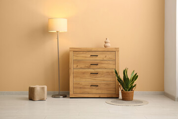 Wooden chest of drawers with houseplant and glowing floor lamp near beige wall in modern room