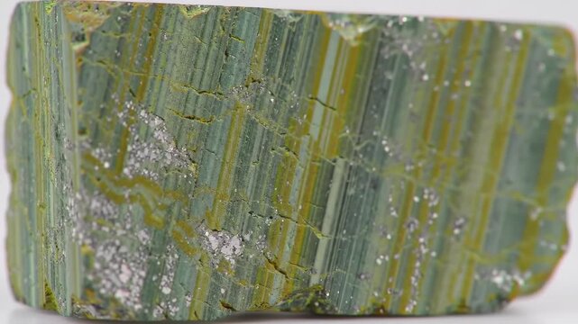 A close up shot of an iridescent green and gold mineral chunk adorned with silver flecks, set against a clean white backdrop. The mineral's texture and pattern is highly detailed.