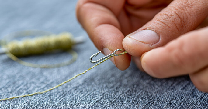 Close-up of hands tying fishing line to a small metal fishing hook on textured fabric surface with spool of line in background