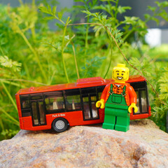 Naklejka premium Lego minifigure with a red bus on a rock in front of green foliage