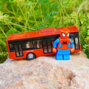 Spider-Man toy figure stands next to a red toy bus on a rock outdoors
