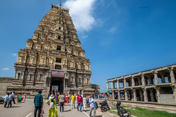 Hampi, India - 01 November 2025: View of the towering Virupaksha Temple against a bright blue sky, with tourists strolling along the paved path, showcasing the architectural grandeur.