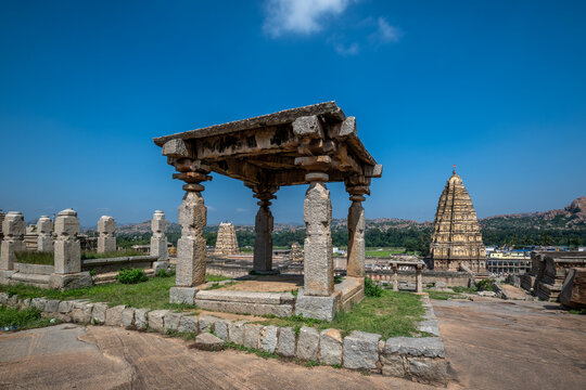 Hampi, India - 01 November 2025: View of the ancient pavilion's weathered stone against the bright sky, with the towering Virupaksha Temple piercing the horizon.