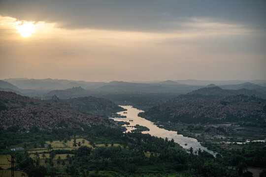 Hampi, India - 02 November 2025: View of the Tungabhadra River winding through a landscape of ancient ruins and lush greenery under a soft, diffused sunset.