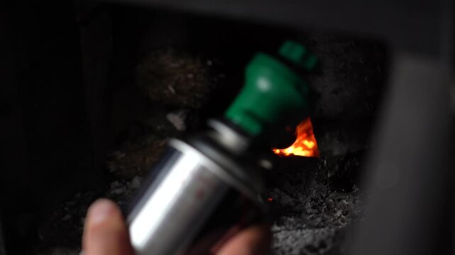 Hand uses green-capped gas igniter to kindle bright embers, fueling small, intense fire in dark stove hearth.