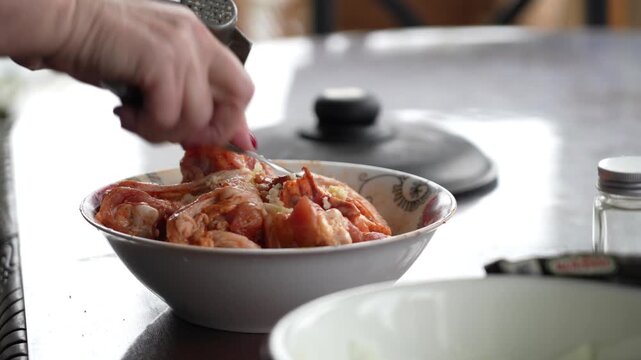 Hand seasons raw chicken cuts with garlic and spices in patterned bowl, preparing meat for cooking.