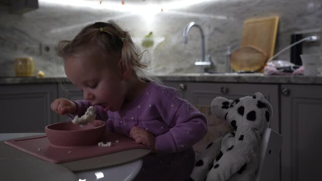 Toddler's independent mealtime in home kitchen, happily self-feeding with spoon, often creating sweet mess, always accompanied by beloved plush pal.