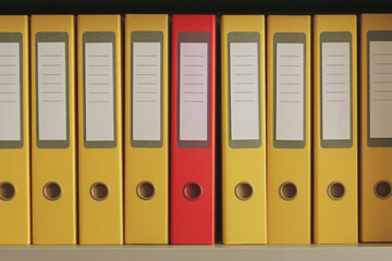 A red folder among yellow ones on a shelf.