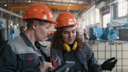 Young female worker of metal fabrication factory in hard hat explaining production progress to middle aged supervisor taking notes on digital tablet during quality control inspection in workshop - Powered by Adobe