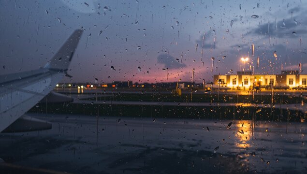 Wing of airplane blurred by raindrops on window during landing in twilight, showcasing the rainy evening atmosphere