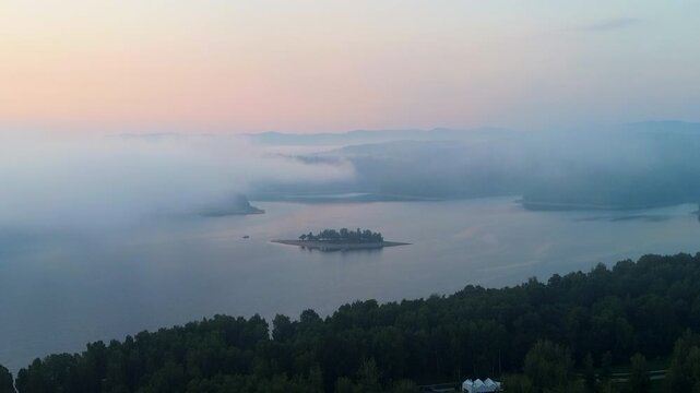 Aerial shot of small forested island surrounded by mist over a calm lake at dawn with soft pastel sky tones. Distant mountain silhouettes. Solina, Poland.