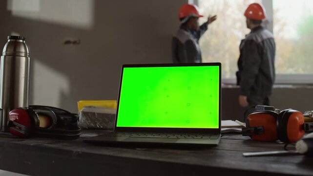 Open laptop with green screen placed on construction table among tools and safety gear while two workers in helmets discussing project plans in background at unfinished building