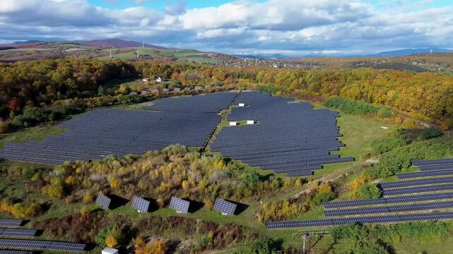 Wide aerial view of a massive solar power plant nestled in an autumn forest. Large field of photovoltaic panels surrounded by colorful trees and hills.