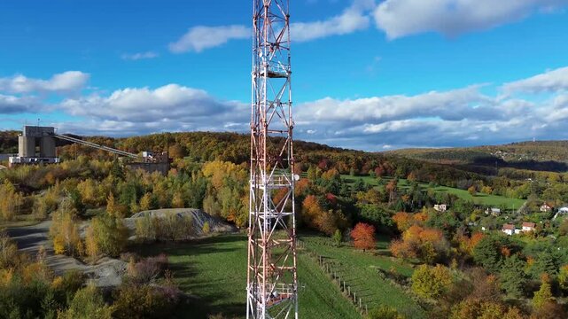Aerial view of a tall red and white telecommunication tower. Steel lattice mast with antennas standing near an abandoned factory in autumn nature.