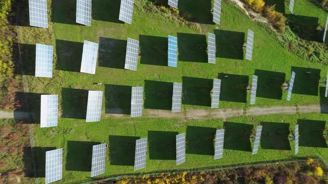 Top-down aerial view of solar panel trackers on a green field. Geometric pattern of photovoltaic modules casting shadows on grass in autumn.