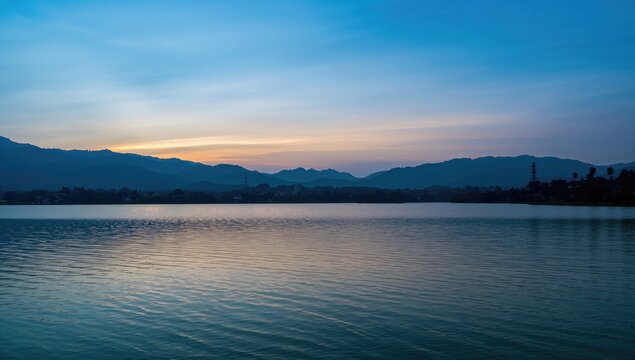 The serene waters of a tranquil lake glow warmly as the sun sets behind distant mountains