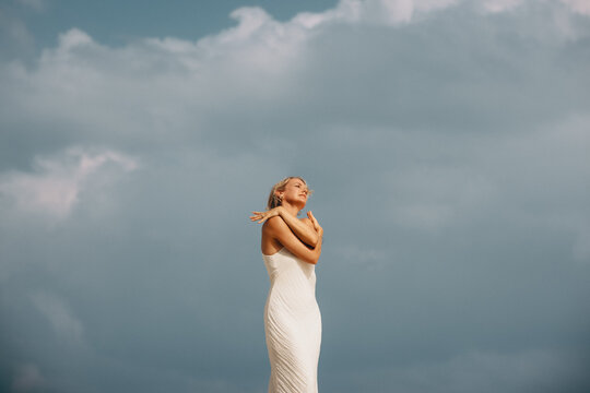 Joyful woman in white dress embracing herself under a moody cloudy sky