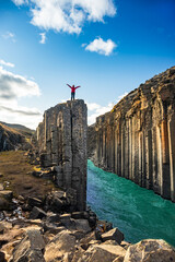 A person stands on rocks in Studlagil Basalt Canyon, surrounded by towering basalt columns and a flowing river in Iceland. The vibrant water contrasts with the rugged landscape.