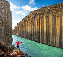 A person stands with arms outstretched on rocky terrain in Studlagil Canyon, Iceland. The striking hexagonal basalt columns frame a vibrant turquoise river, showcasing natural beauty