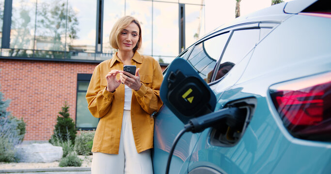 Woman using smartphone app while standing next to her electric car charging at a portable charging station on a city street.