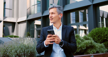 Businessman using smartphone outdoors standing on city street, smiling looking to mobile phone screen, enjoying urban atmosphere. Cheerful male using mobile application, texting message, shopping