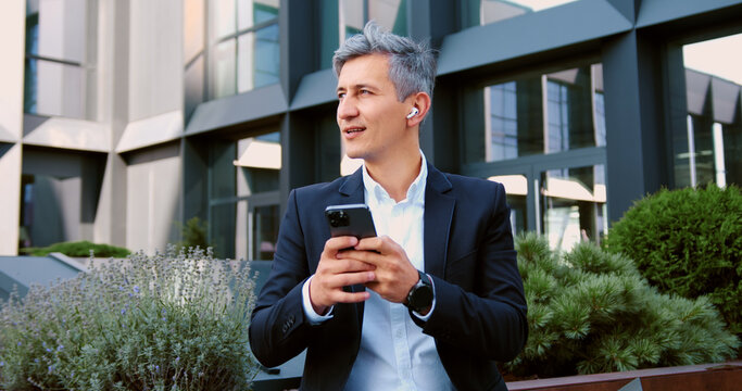 Businessman using smartphone outdoors standing on city street, smiling looking to mobile phone screen, enjoying urban atmosphere. Cheerful male using mobile application, texting message, shopping