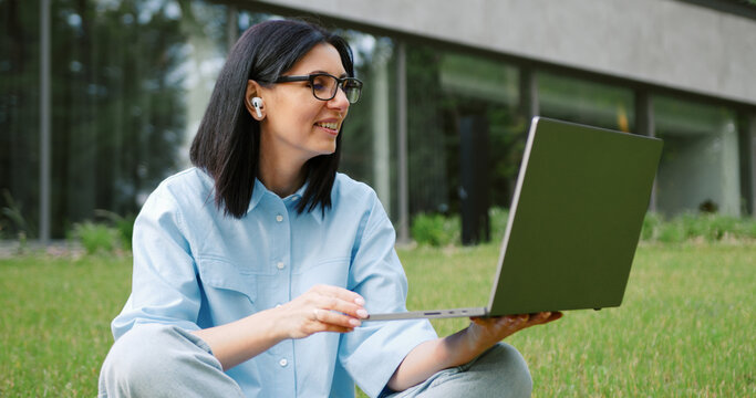 Close-up of a young brunette woman sitting on the grass in a city park, wearing headphones, participating in an online business meeting or e-learning session on her laptop