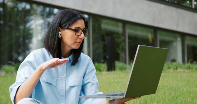 Close up of freelance designer sitting cross-legged on the grass with headphones, editing a creative project on her laptop in a relaxed park setting