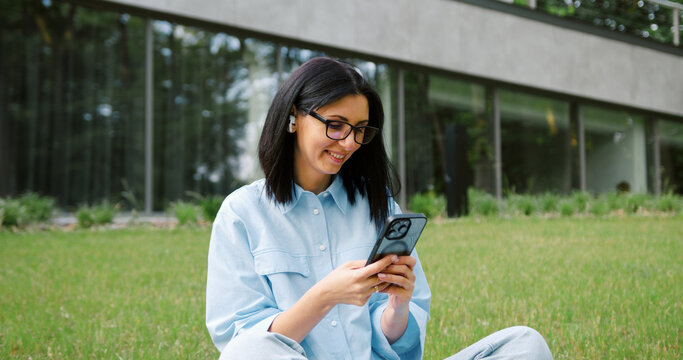 Attractive young woman with a beautiful smile sitting cross-legged on the grass in a green park, using her smartphone, enjoying a sunny day