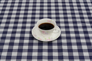 Top view of a porcelain coffee cup with floral pattern and espresso, on a matching saucer. Set on a large, navy blue and white plaid tablecloth. Symbolizes breakfast, break, and home.