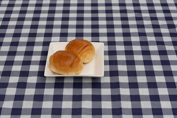 Two sweet rolls on a white square plate, centered on a navy blue and white plaid tablecloth. Top view. Symbolizes breakfast, bakery, snack, and simple comfort food.