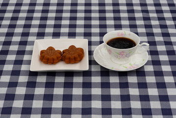 Top view of a coffee cup and a plate with two maple leaf-shaped pastries on a navy blue and white plaid tablecloth. Ideal for breakfast or autumn snack concept.