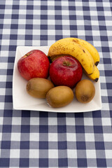 Close-up of a white square plate with fresh fruit (apples, kiwis, bananas) on a navy blue and white plaid tablecloth. Vertical orientation. Ideal for healthy snack and diet concepts.