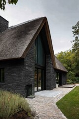 Fototapeta premium A modern black stone house adorned with a thatched roof sits amid a lush green landscape, creating a tranquil atmosphere as dusk falls