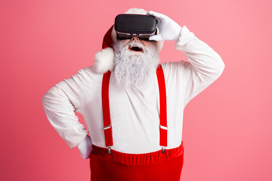 Santa in a red suit with suspenders wears a virtual reality headset and smiles against a pink background creating a festive moment