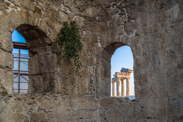 An ancient Roman wall with arched window framing the Temple of Apollo with Corinthian columns, adorned with green ivy vines. Historic architecture in Side, Antalya, Turkey.

