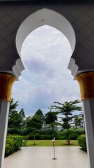 Scenic view through a mosque arch revealing a green garden and cloudy sky. A peaceful composition uniting Islamic architecture and nature’s calm beauty.
