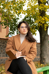 Young attractive woman in brown coat sitting on bench and writing in notebook