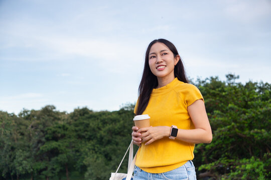 Happy Asian woman smiling, holding a cup of coffee or tea outdoors in a park or natural area