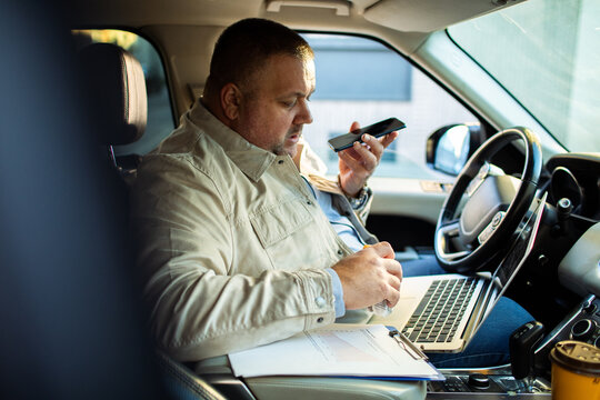 Adult man focused, multitasking with laptop and phone in parked car
