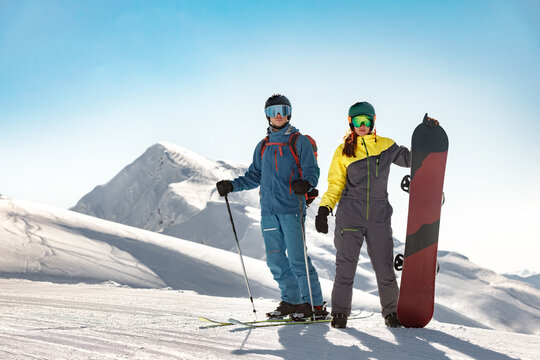 Couple skier and snowboarder stand at the top of a ski resort slope on sunny day with snow-capped peaks in the background.