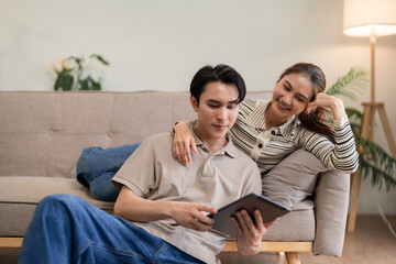 Asian adult couple enjoying digital content on tablet relaxing together at cozy home living room partners bonding modern lifestyle