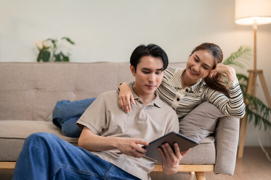 Asian adult couple enjoying digital content on tablet relaxing together at cozy home living room partners bonding modern lifestyle
