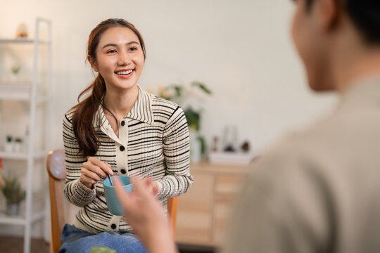 Happy Asian adult couple engaging cheerful conversation enjoying morning coffee or tea at home in cozy living room connecting relationship bonding