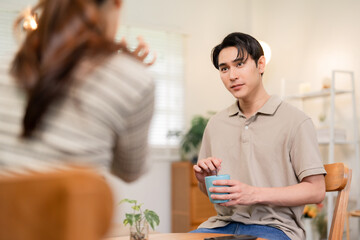Young Asian adult man enjoys coffee drink conversation partner home cozy living room setting. Relaxed moments sharing domestic life comfort.