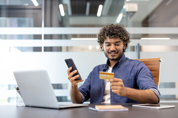 Smiling young Indian man sitting at office desk, using mobile phone and credit card
