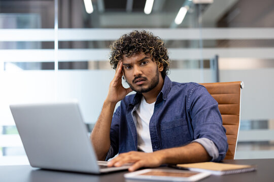 Portrait of a young shocked Indian man sitting at a desk in the office with a laptop, holding his head in frustration and looking at the camera