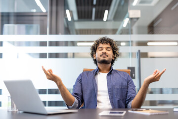 Relaxed Indian young man sitting in the office at his desk with his eyes closed in the lotus position and meditating